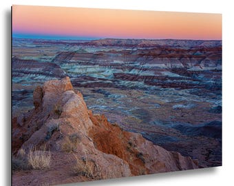 May include: A landscape photograph of a desert badlands terrain featuring layered sedimentary rock formations in shades of red, orange, and blue. The horizon is illuminated by a soft sunset sky with gradients of pink, orange, and deep blue.
