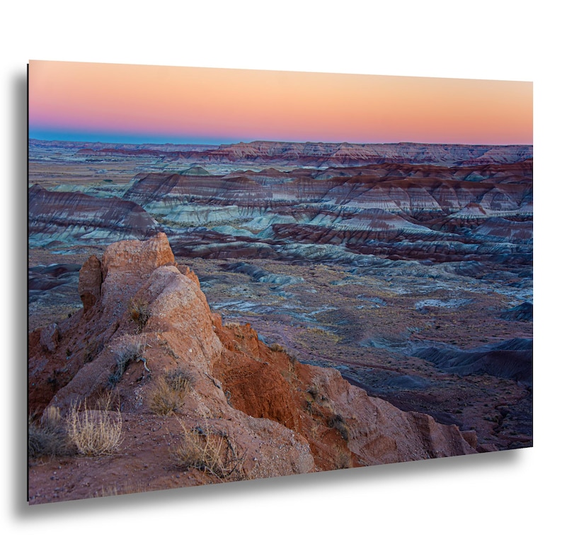 May include: A landscape photograph of a desert badlands terrain featuring layered sedimentary rock formations in shades of red, orange, and blue. The horizon is illuminated by a soft sunset sky with gradients of pink, orange, and deep blue.