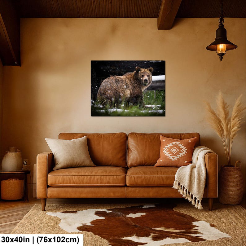 May include: A rectangular wall art print featuring a brown bear standing in a grassy field during a light snowfall. The artwork measures 76 by 102 cm and is displayed in a living room setting above a brown leather sofa with pillows.