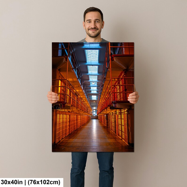 May include: A rectangular wall art print measuring 30 by 40 inches, featuring a perspective view of a prison cell block. The image displays rows of orange-toned metal cells on both sides, a central walkway, and a ceiling with blue-lit skylights.