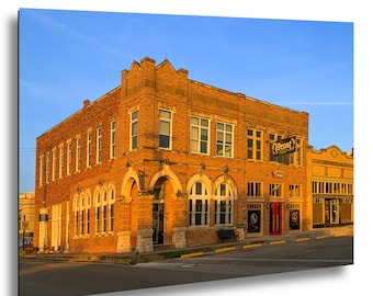 May include: A two-story brick building on a street corner featuring arched windows on the ground floor, rectangular windows on the second floor, and a sign reading Prost. The exterior is illuminated by warm golden sunlight against a clear blue sky.