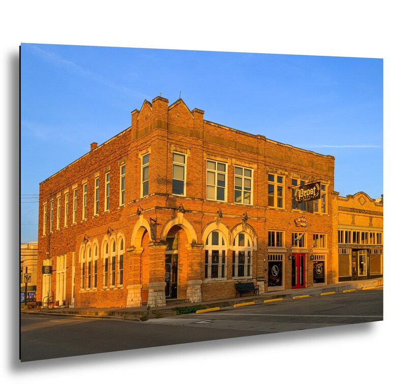 May include: A two-story brick building on a street corner featuring arched windows on the ground floor, rectangular windows on the second floor, and a sign reading Prost. The exterior is illuminated by warm golden sunlight against a clear blue sky.