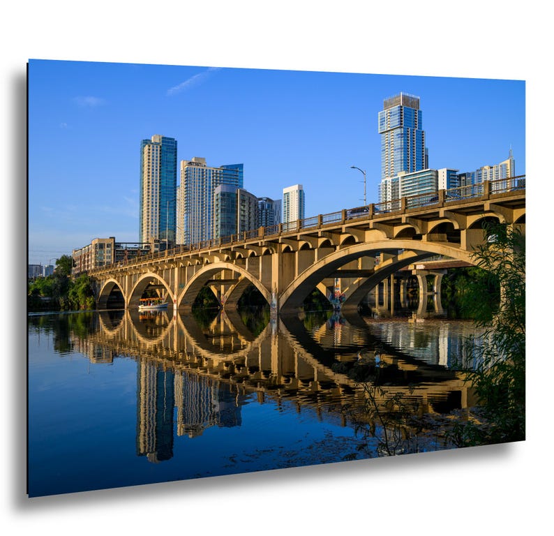 Lamar Boulevard Bridge and the downtown Austin skyline reflected in calm Lady Bird Lake. Sunlit concrete arches form nearly complete circles with their reflections beneath a clear blue sky. A boat passes under the center of one arch. A concrete arch bridge spans a calm river under a clear blue sky, reflecting in the water below. In the background, a cluster of tall glass skyscrapers rises above the horizon, illuminated by the warm light of the late afternoon sun.  Photography print.