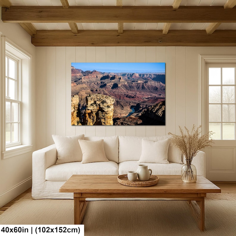 May include: A rectangular wall art print depicting a panoramic view of the Grand Canyon with layered red rock formations and a winding river below, displayed in a living room above a white sofa and a wooden coffee table, measuring 40x60 inches.