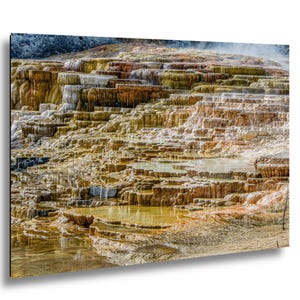 Könnte beinhalten: Eine Landschaftsaufnahme der Mammoth Hot Springs mit gestuften Travertinterrassen und fließendem Wasser. Die Mineralablagerungen zeigen Schattierungen in Orange, Gelb, Braun und Weiß und bilden eine geschichtete, terrassenförmige geologische Formation.
