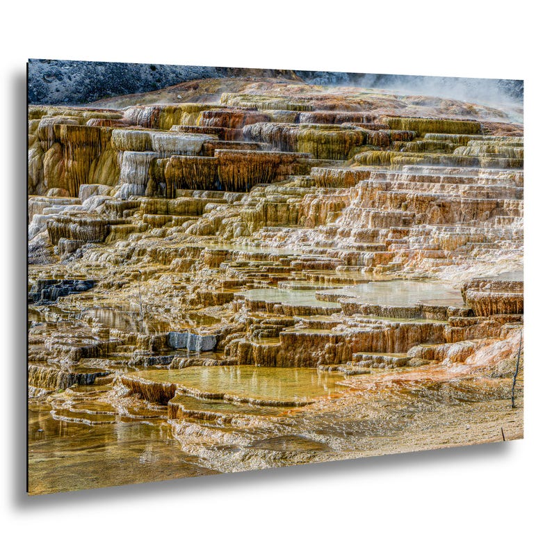 May include: A landscape photograph of Mammoth Hot Springs featuring tiered travertine terraces with flowing water. The mineral deposits display shades of orange, yellow, brown, and white, creating a layered, stepped geological formation.