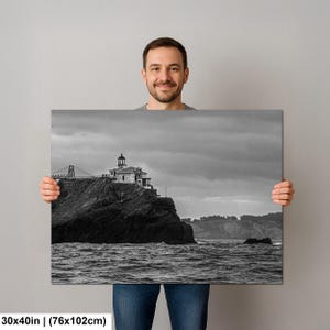 May include: A rectangular wall art print measuring 30 by 40 inches featuring a black and white landscape photograph of a lighthouse perched on a rocky cliffside above a turbulent ocean with a cloudy sky and a bridge structure in the distance.