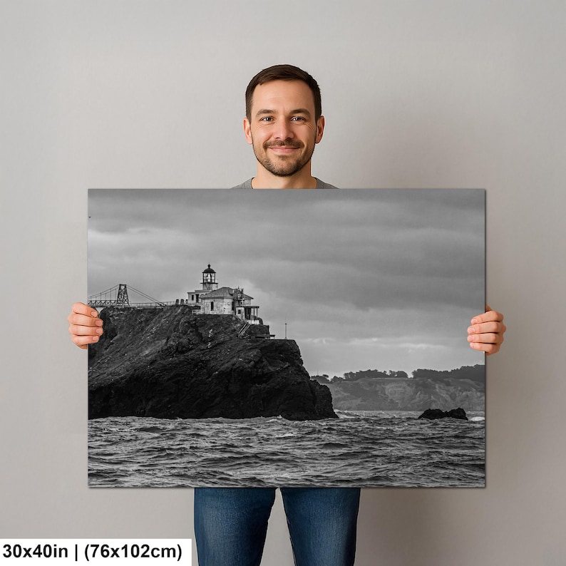 May include: A rectangular wall art print measuring 30 by 40 inches featuring a black and white landscape photograph of a lighthouse perched on a rocky cliffside above a turbulent ocean with a cloudy sky and a bridge structure in the distance.