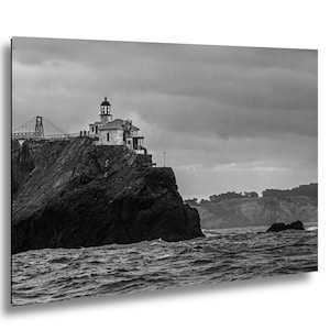 May include: A black and white photograph of a lighthouse perched on a rugged, dark cliffside overlooking a turbulent ocean. A suspension bridge structure is visible in the background, and the sky is filled with heavy, overcast clouds.