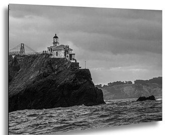 May include: A black and white photograph of a lighthouse perched on a rugged, dark cliffside overlooking a turbulent ocean. A suspension bridge structure is visible in the background, and the sky is filled with heavy, overcast clouds.