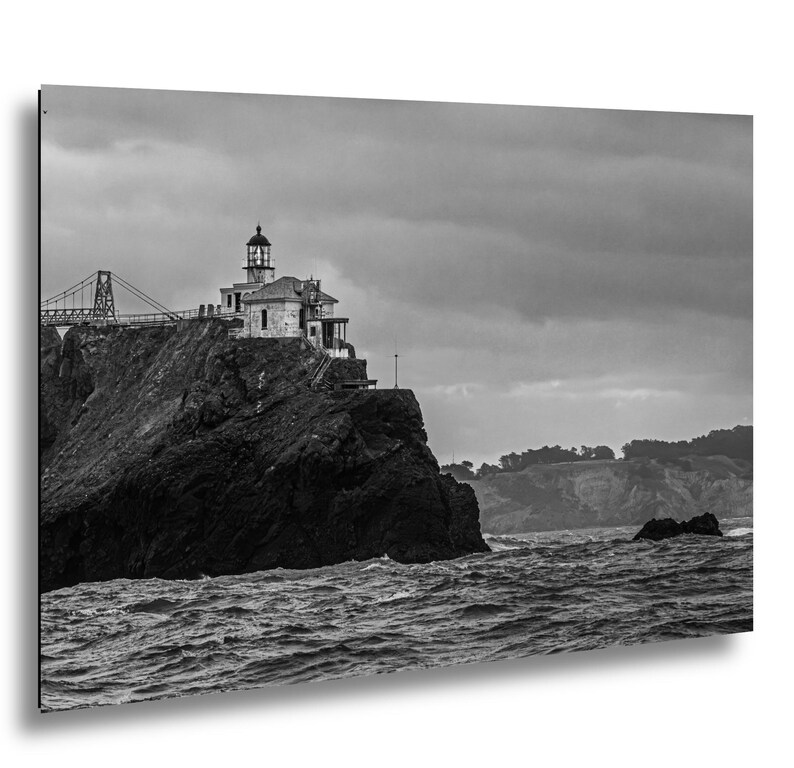 May include: A black and white photograph of a lighthouse perched on a rugged, dark cliffside overlooking a turbulent ocean. A suspension bridge structure is visible in the background, and the sky is filled with heavy, overcast clouds.