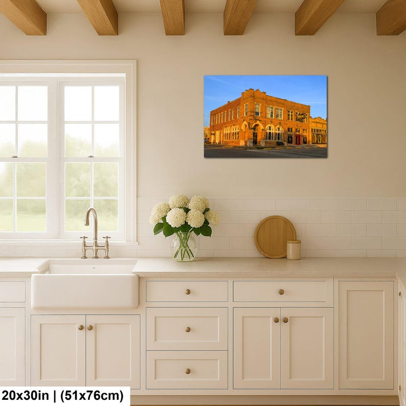 May include: A rectangular wall art print featuring a multi-story brick building illuminated by warm golden sunlight against a clear blue sky. The print is displayed on a wall above a white kitchen counter with a sink, faucet, and flowers.