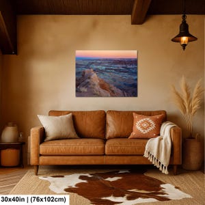 May include: A rectangular wall art print featuring a desert landscape at sunset with layered rock formations in shades of blue, purple, and orange. The print measures 30 inches by 40 inches and is displayed on a plain wall above a leather sofa.