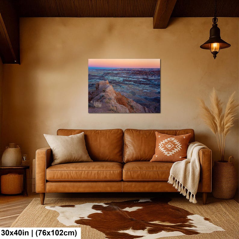 May include: A rectangular wall art print featuring a desert landscape at sunset with layered rock formations in shades of blue, purple, and orange. The print measures 30 inches by 40 inches and is displayed on a plain wall above a leather sofa.