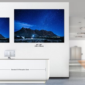 Puede incluir: Dos impresiones de arte mural rectangulares que muestran un paisaje nocturno con una cordillera bajo un cielo azul estrellado. La impresi&oacute;n peque&ntilde;a mide 76 x 102 cm y la grande 102 x 152 cm, expuestas sobre un mostrador de recepci&oacute;n de 5 pies.
