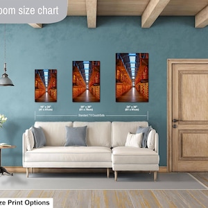 May include: A living room size chart displaying three rectangular wall art prints of a warehouse interior with orange lighting. The prints are labeled with dimensions 16" x 24", 20" x 30", and 24" x 36" above a white sectional sofa and a small side table.