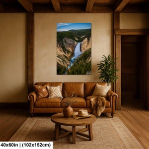 May include: A rectangular wall art print measuring 40x60 inches featuring a landscape photograph of a waterfall cascading through a deep canyon with pine trees and a blue sky. The print is displayed on a beige wall above a brown leather sofa.
