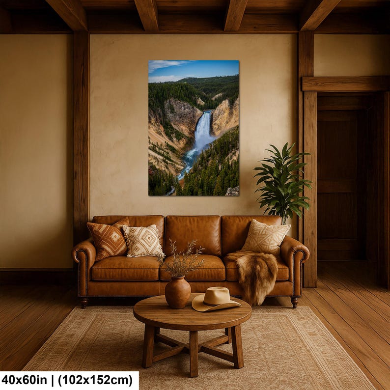 May include: A rectangular wall art print measuring 40x60 inches featuring a landscape photograph of a waterfall cascading through a deep canyon with pine trees and a blue sky. The print is displayed on a beige wall above a brown leather sofa.