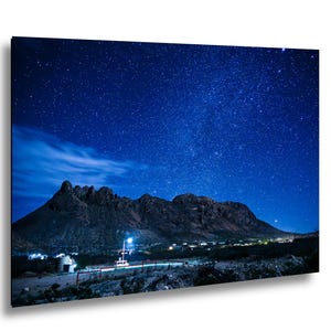 May include: A rectangular wall art print featuring a night landscape with a dark mountain range silhouetted against a deep blue sky filled with numerous bright stars. A small cluster of illuminated buildings sits at the base of the mountains.