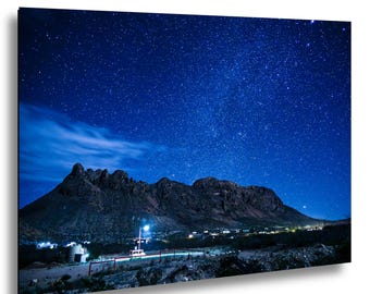 May include: A rectangular wall art print featuring a night landscape with a dark mountain range silhouetted against a deep blue sky filled with numerous bright stars. A small cluster of illuminated buildings sits at the base of the mountains.