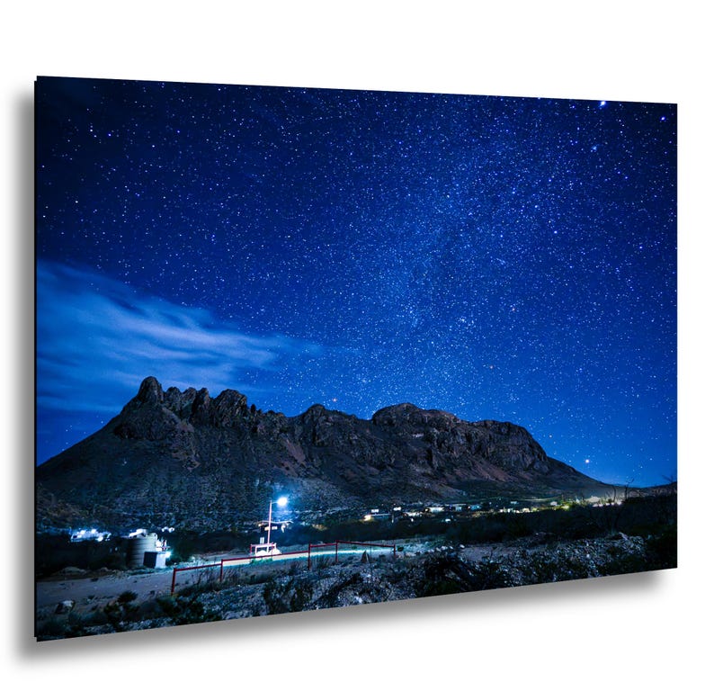 Puede incluir: Un cuadro rectangular que muestra un paisaje nocturno con una cordillera oscura silueteada contra un cielo azul profundo lleno de numerosas estrellas brillantes. Un peque&ntilde;o grupo de edificios iluminados se asienta en la base.