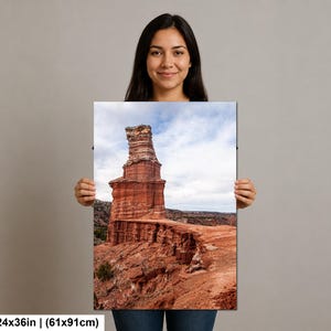 May include: A rectangular wall art print measuring 24 by 36 inches, featuring a landscape photograph of a tall, reddish-brown rock formation rising from a canyon floor under a cloudy sky, with visible layered geological textures and earthy tones.
