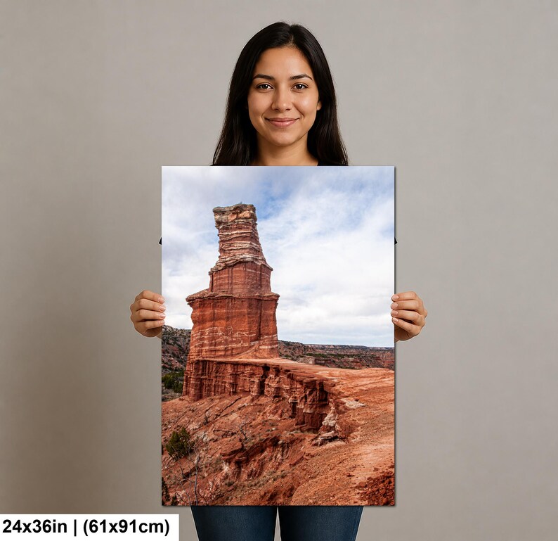 May include: A rectangular wall art print measuring 24 by 36 inches, featuring a landscape photograph of a tall, reddish-brown rock formation rising from a canyon floor under a cloudy sky, with visible layered geological textures and earthy tones.