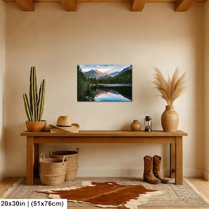 May include: A rectangular wall art print measuring 20 by 30 inches, featuring a landscape photograph of a calm lake reflecting snow-capped mountains at sunrise, surrounded by a dense forest of green pine trees under a soft blue and orange sky.