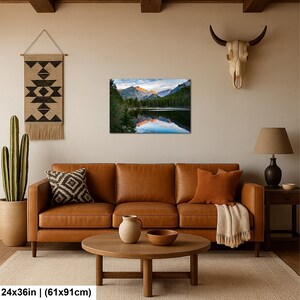 May include: A rectangular wall art print featuring a mountain landscape with a lake, pine trees, and a golden sunrise reflection. The print measures 24 inches by 36 inches and is displayed on a beige wall above a brown leather sofa and coffee table.