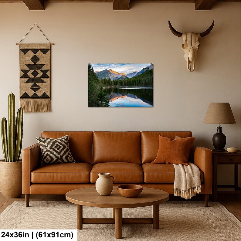 May include: A rectangular wall art print featuring a mountain landscape with a lake, pine trees, and a golden sunrise reflection. The print measures 24 inches by 36 inches and is displayed on a beige wall above a brown leather sofa and coffee table.