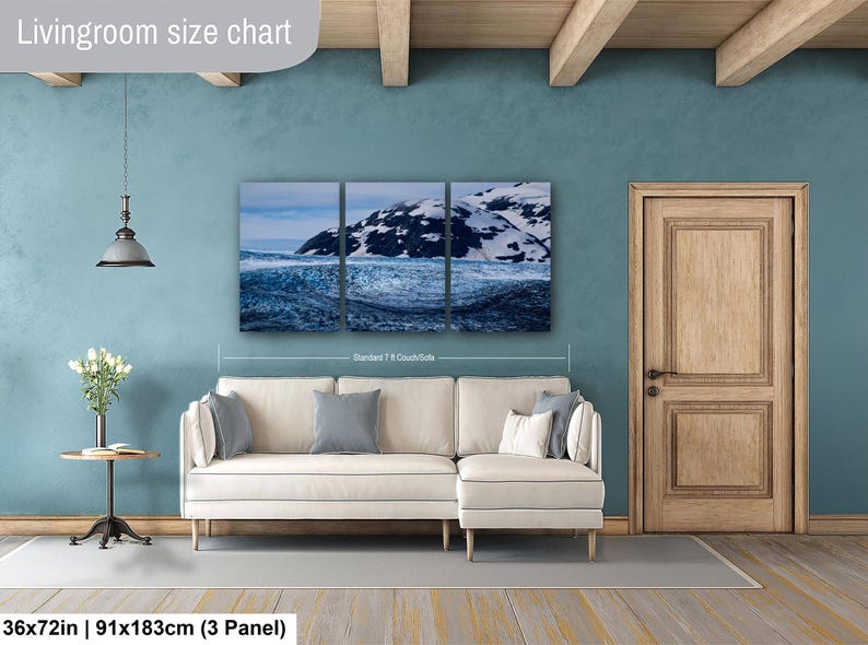 May include: A three-panel wall art set depicting a snowy mountain landscape above a dark blue ocean, displayed above a cream-colored sectional sofa. The text "Livingroom size chart" and "36x72in | 91x183cm (3 Panel)" is visible on the image.