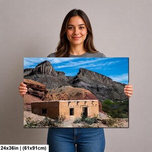 May include: A rectangular wall art print measuring 24 by 36 inches, featuring a landscape photograph of a tan adobe building situated at the base of rugged, dark gray rocky mountains under a bright blue sky with scattered white clouds.