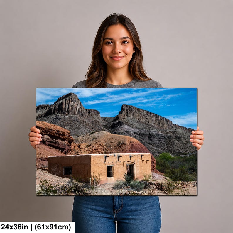 May include: A rectangular wall art print measuring 24 by 36 inches, featuring a landscape photograph of a tan adobe building situated at the base of rugged, dark gray rocky mountains under a bright blue sky with scattered white clouds.