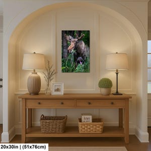 May include: A rectangular wall art print featuring a brown moose standing in a lush green forest, eating leaves. The print measures 20 by 30 inches and is displayed on a wall above a wooden console table with two lamps, a small frame, and baskets.