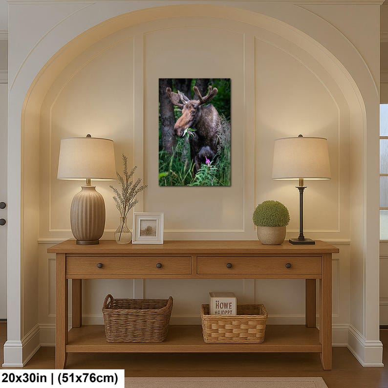 May include: A rectangular wall art print featuring a brown moose standing in a lush green forest, eating leaves. The print measures 20 by 30 inches and is displayed on a wall above a wooden console table with two lamps, a small frame, and baskets.