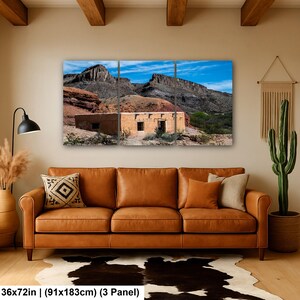 May include: A three-panel wall art set depicting a desert landscape with a tan adobe building nestled against rugged, dark rocky mountains under a bright blue sky. The text at the bottom reads 36x72in (91x183cm) (3 Panel).