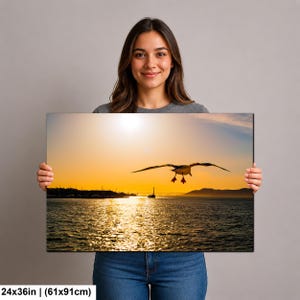 May include: A rectangular wall art print measuring 24 by 36 inches, featuring a photograph of a seagull in flight over a body of water at sunset. The scene includes a distant shoreline, a small boat, and a golden sky reflecting on the water.