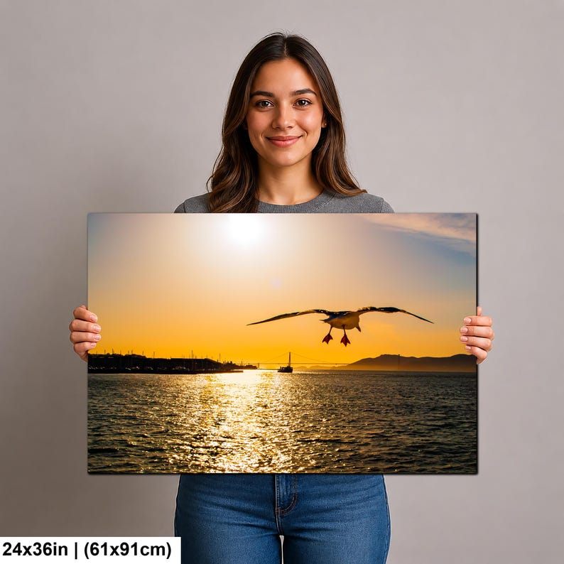 May include: A rectangular wall art print measuring 24 by 36 inches, featuring a photograph of a seagull in flight over a body of water at sunset. The scene includes a distant shoreline, a small boat, and a golden sky reflecting on the water.