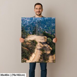 以下が含まれることがあります： 76 x 102 cmの長方形のウォールアートプリント。乾燥した草の生い茂る丘を登る未舗装のハイキングコースと、その先にある2本の背の高い細い木、そして背景に広がる街並みを俯瞰で捉えた風景写真が印刷されています。