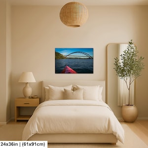 May include: A rectangular wall art print measuring 24 inches by 36 inches, featuring a landscape photograph of a red kayak bow on dark blue water, looking toward a large arched bridge under a bright blue sky with light clouds.