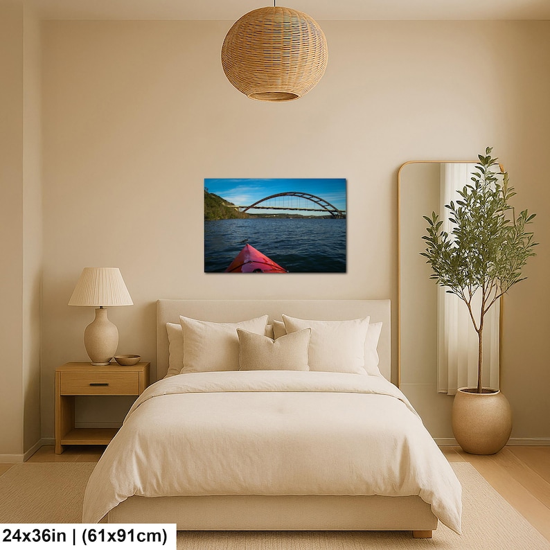May include: A rectangular wall art print measuring 24 inches by 36 inches, featuring a landscape photograph of a red kayak bow on dark blue water, looking toward a large arched bridge under a bright blue sky with light clouds.