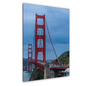 May include: A vertical wall art print featuring the Golden Gate Bridge in San Francisco. The iconic bright orange-red suspension bridge spans across dark blue water under a cloudy sky, with green hills visible on the sides of the structure.