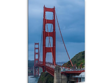 May include: A vertical wall art print featuring the Golden Gate Bridge in San Francisco. The iconic bright orange-red suspension bridge spans across dark blue water under a cloudy sky, with green hills visible on the sides of the structure.
