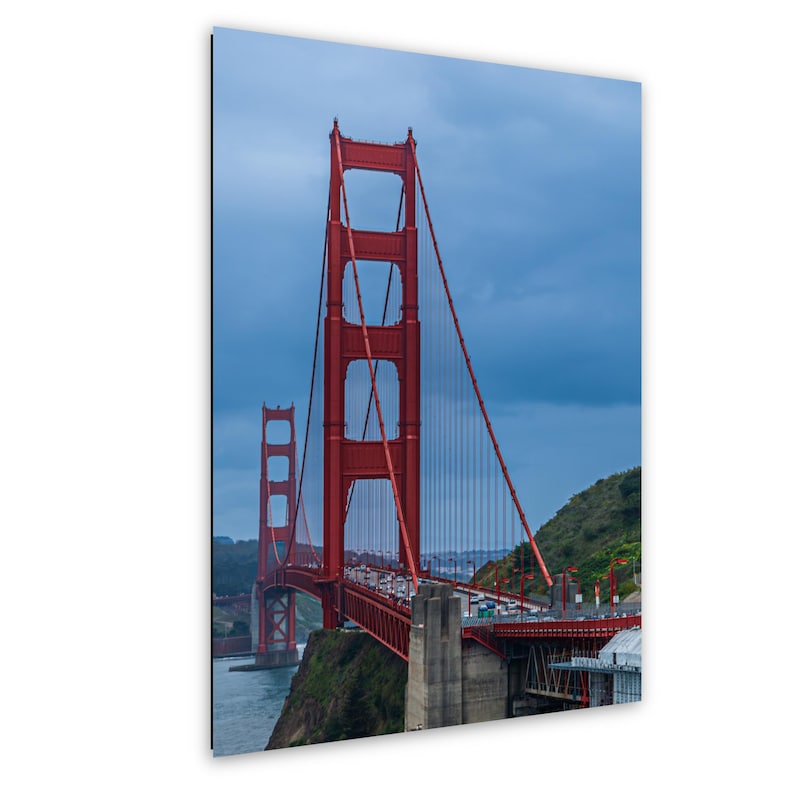 May include: A vertical wall art print featuring the Golden Gate Bridge in San Francisco. The iconic bright orange-red suspension bridge spans across dark blue water under a cloudy sky, with green hills visible on the sides of the structure.