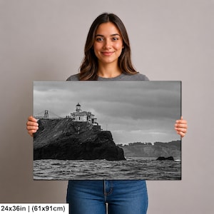 May include: A rectangular wall art print measuring 24 by 36 inches, featuring a black and white landscape photograph of a lighthouse perched on a rocky cliffside above the ocean, with a bridge structure visible in the distant background.