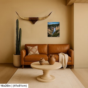 May include: A rectangular wall art print depicting a waterfall in a canyon, displayed above a tan leather sofa. The scene includes a tall cactus in a textured pot, a patterned throw pillow, two ceramic vases on a round table, and long horns.