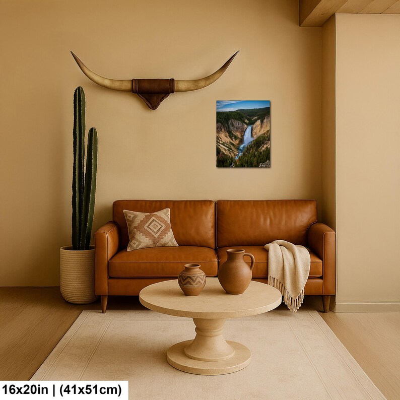 May include: A rectangular wall art print depicting a waterfall in a canyon, displayed above a tan leather sofa. The scene includes a tall cactus in a textured pot, a patterned throw pillow, two ceramic vases on a round table, and long horns.