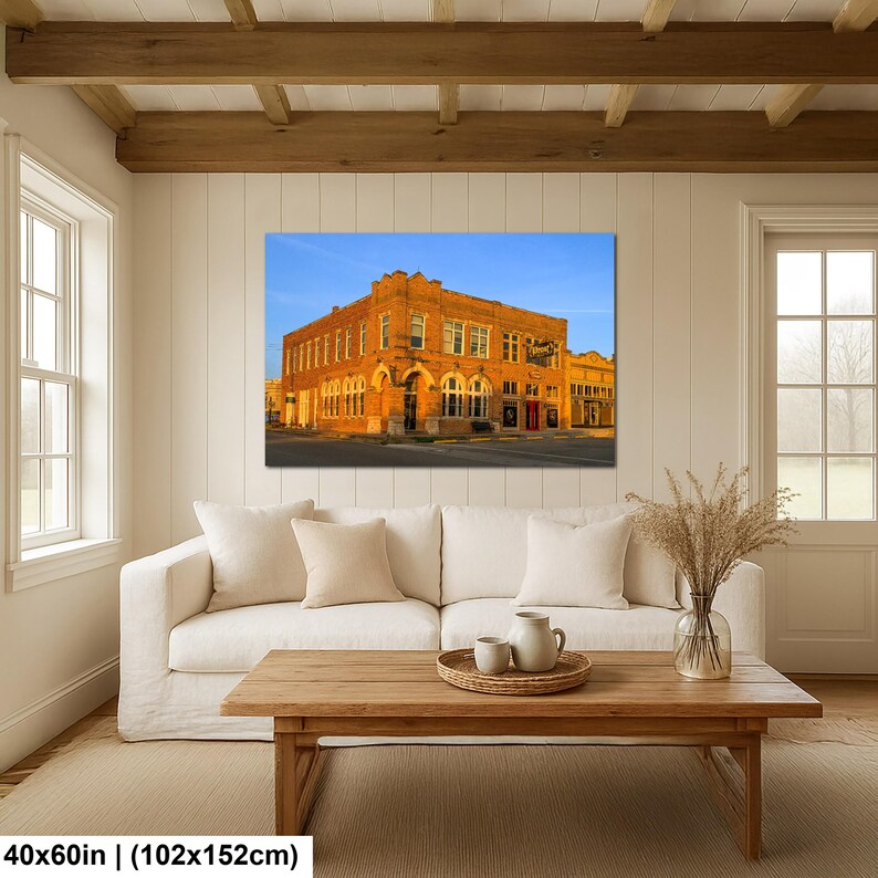 May include: A rectangular wall art print featuring a multi-story brick building at sunset, displayed above a white sofa and wooden coffee table. The print measures 40x60 inches and depicts a corner structure with arched windows and warm lighting.