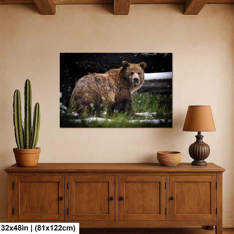 May include: A rectangular wall art print featuring a brown grizzly bear standing in a snowy, grassy landscape. The bear has thick, textured fur and is positioned in front of a dark, horizontal log. The print measures 81 by 122 cm.