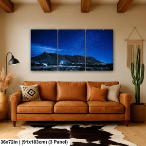 Puede incluir: Un conjunto de arte mural de tres paneles que representa un paisaje nocturno con una cordillera bajo un cielo azul estrellado. La obra mide 91x183 cm y se exhibe sobre un sof&aacute; de cuero beige con cojines decorativos y una alfombra de piel de vaca.